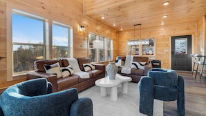 A spacious living area with wooden walls and large windows, furnished with leather sofas and blue chairs, and a central white coffee table. - VIEWS FROM THE MOUNTAIN TOP is Ready for Company (SEVIERVILLE)