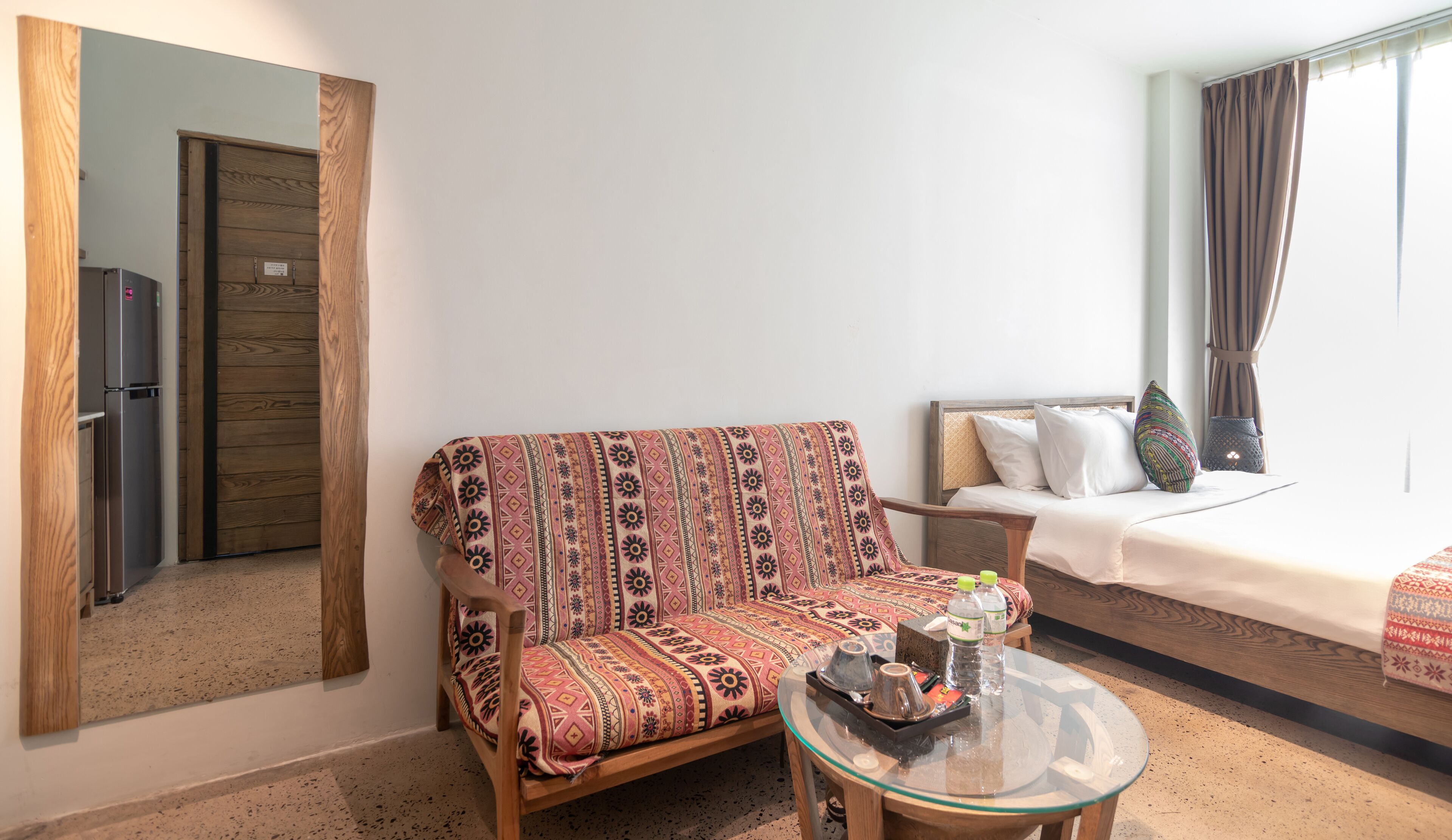 Quarto de hotel aconchegante com sofá de madeira, mesa de centro de vidro, cama com colcha estampada e um espelho grande na parede.