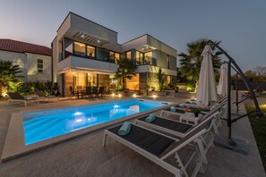 Et moderne hus med to etager, en swimmingpool, udendørs siddepladser og parasoller.