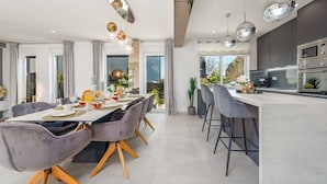 Une cuisine moderne dotée d’appareils électroménagers en acier inoxydable, un coin repas avec une table blanche et des chaises grises, et une grande fenêtre qui laisse entrer la lumière naturelle.