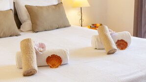 A neatly made bed with rolled towels and a seashell, a lamp on the nightstand, and a view of a window with curtains. - Villa Bellegarde (Albufeira)