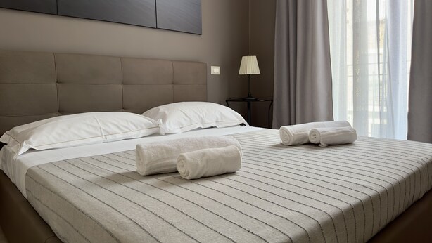 A neatly made bed with two pillows and two rolled towels, a bedside lamp, and a large window with curtains. - Spacious apartment within walking distance of Duomo and Pescheria (Catania)