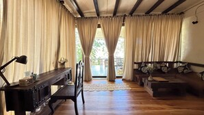 A room with a wooden desk, a chair, a sofa, and a view of a balcony through large curtains.