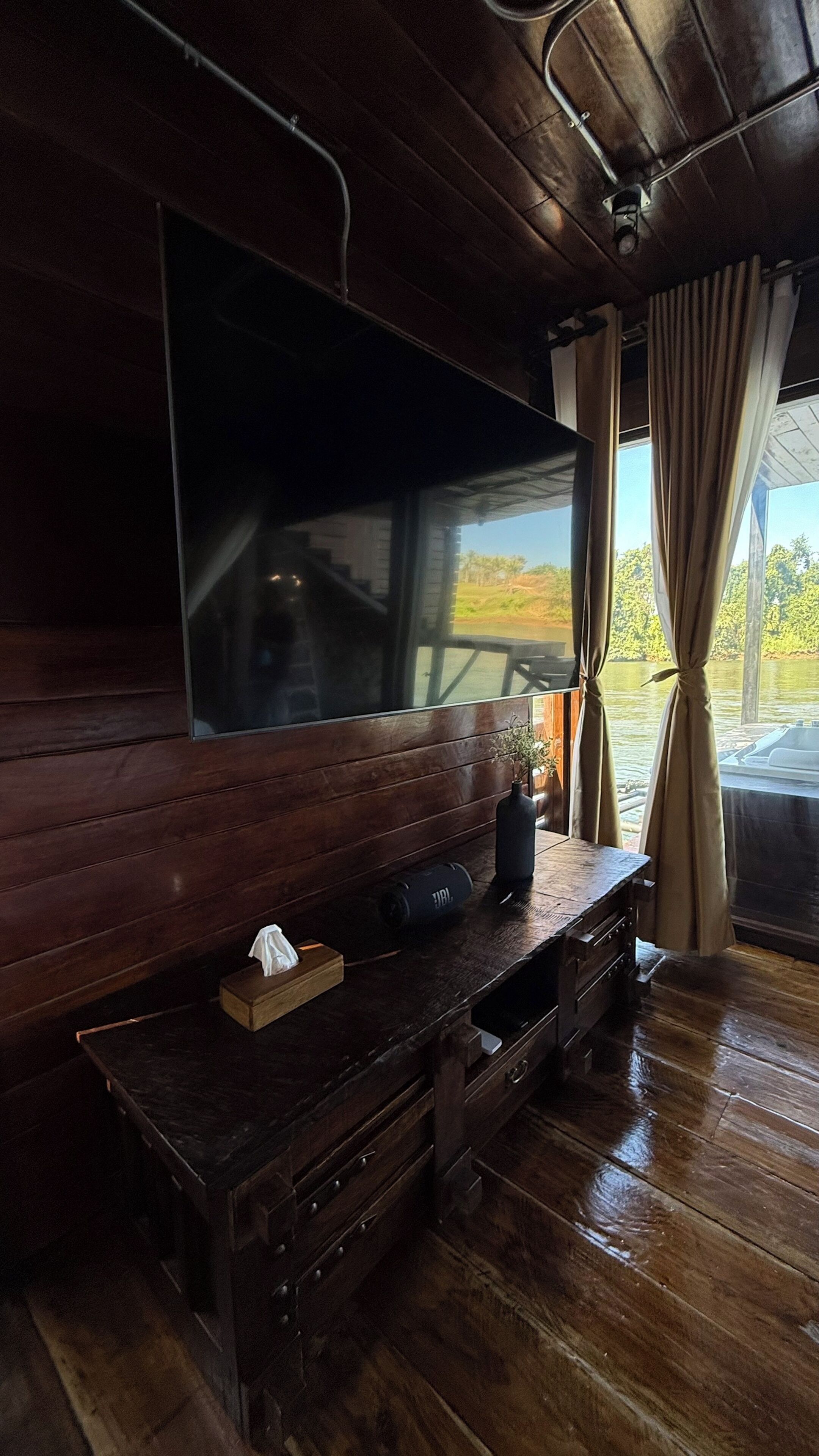 A wooden interior with a large flat-screen TV mounted on the wall, a wooden bench, a small table with a tissue box, and a window with curtains.
