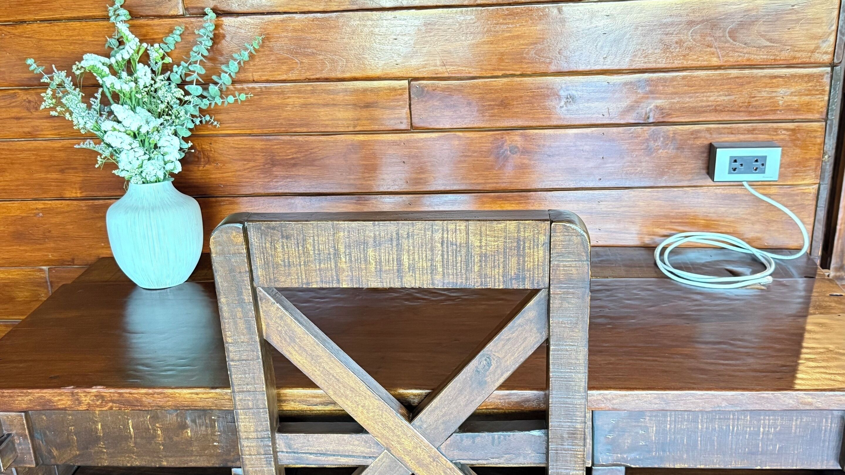A wooden bench with a cross-back design, a vase with green foliage, and a power outlet on the wall.