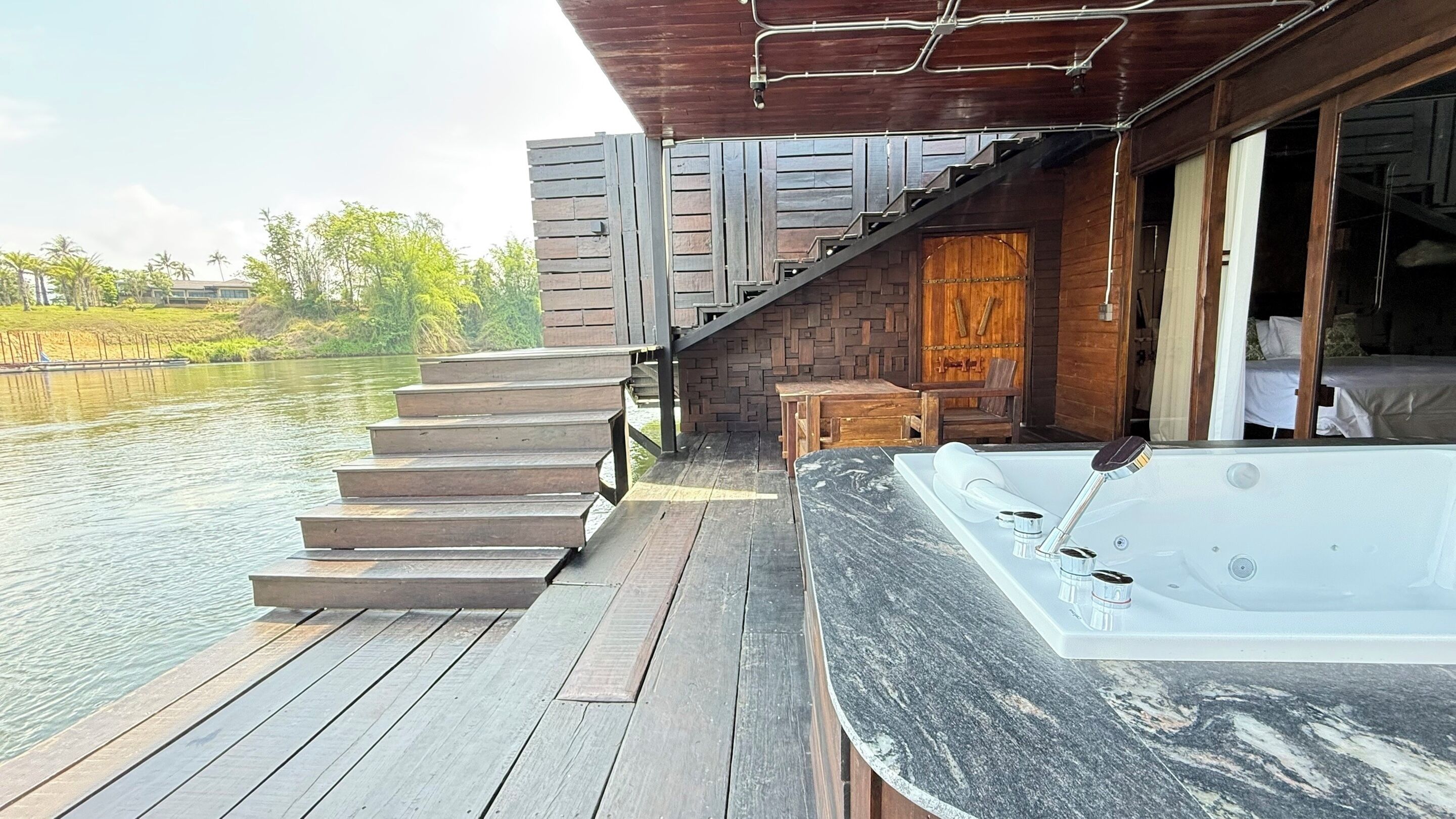 Une terrasse en bois avec un jacuzzi, un escalier menant à une porte en bois et une vue sur une étendue d’eau calme.