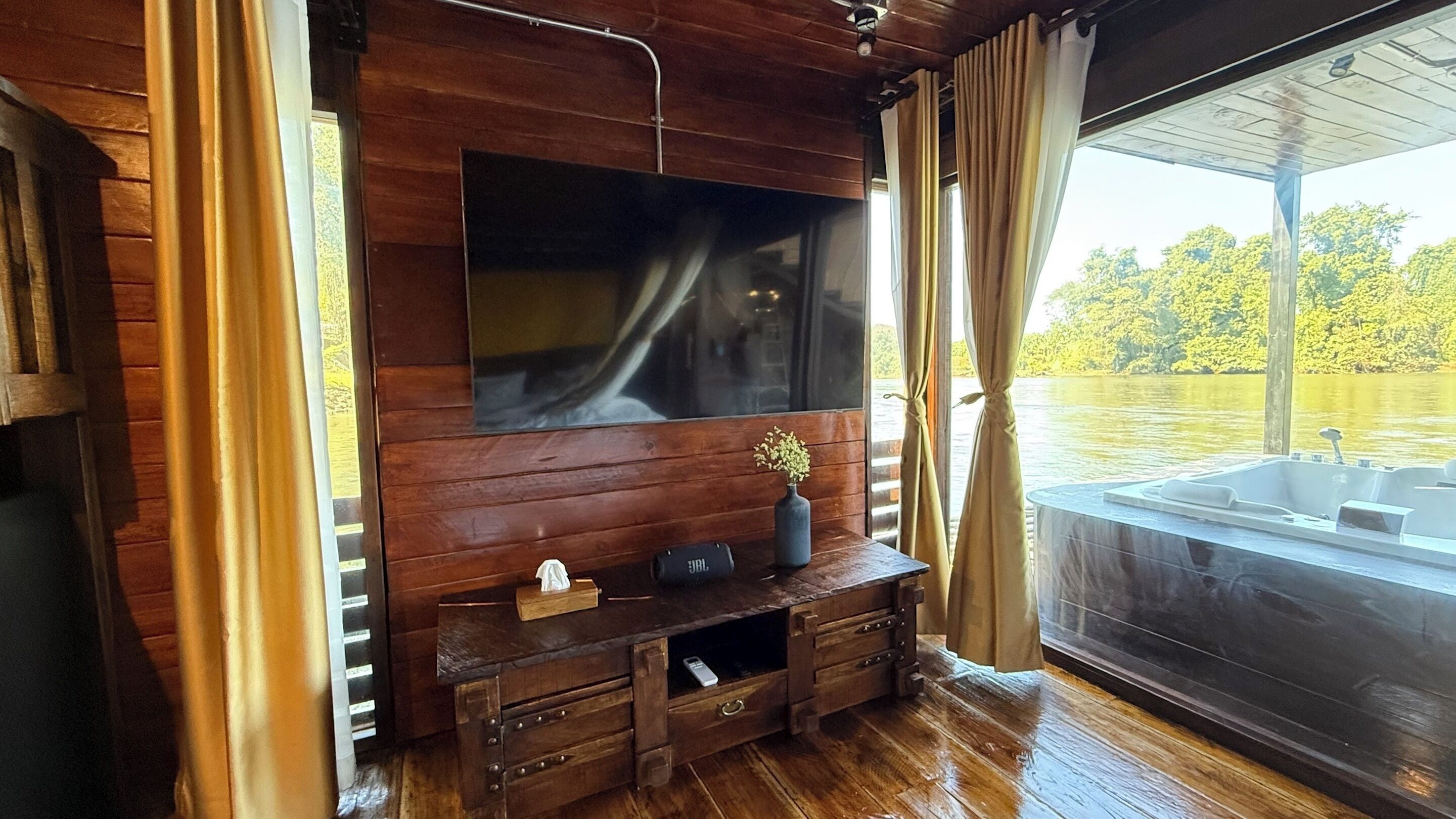 A wooden interior with a large flat-screen TV mounted on the wall, a wooden bench with a vase of flowers, and a view of a lake through large windows.