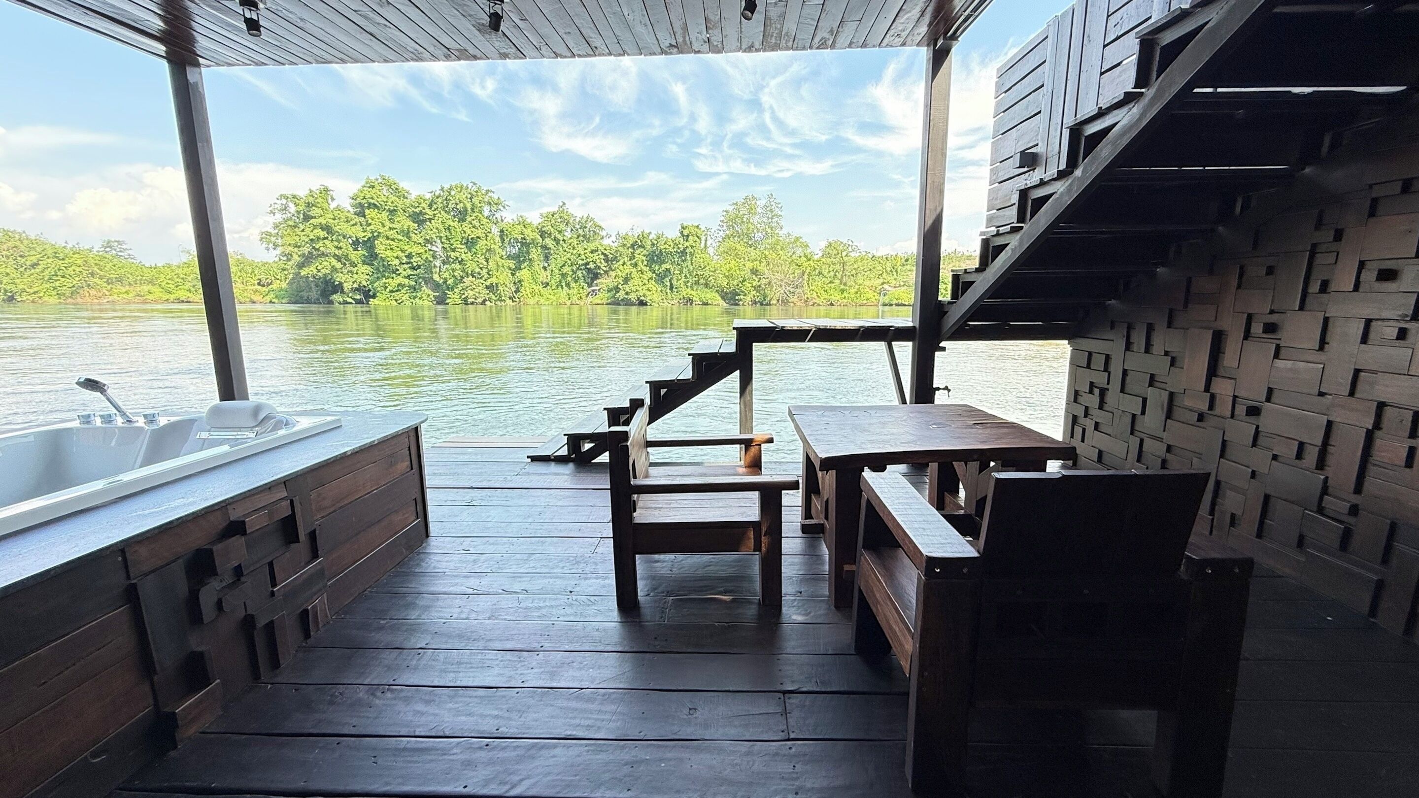 A wooden deck with a hot tub, wooden benches, and a table overlooking a calm lake with trees in the background.