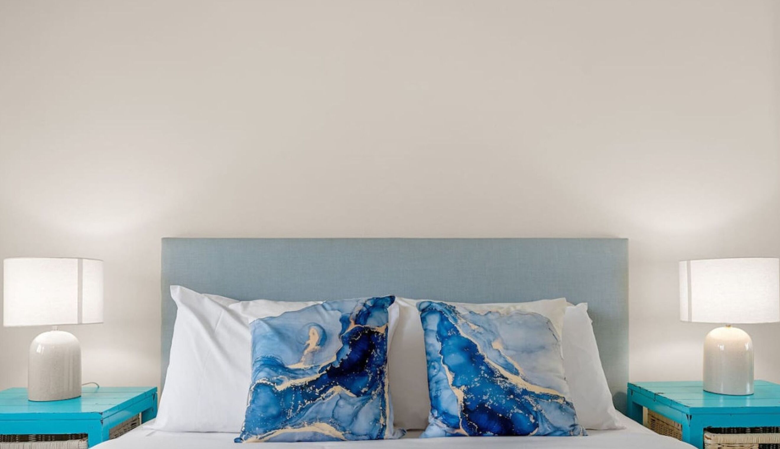 A bed with a light blue headboard, two white pillows, and two bedside tables with white lamps and teal storage boxes.