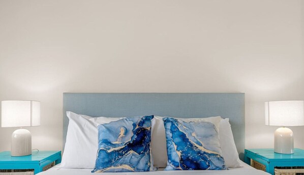 Uma cama com cabeceira azul clara, duas almofadas brancas e duas mesas de cabeceira com luminárias brancas e caixas organizadoras azul-esverdeadas.