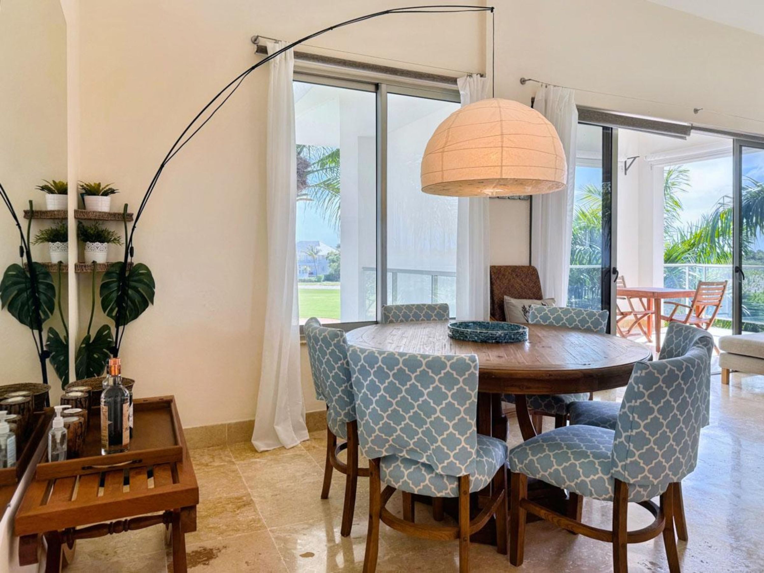 A dining area with a round table, patterned chairs, a hanging lamp, and a view of the outdoors through large windows.
