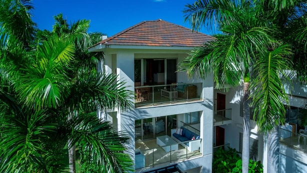 Une maison moderne à deux étages, dotée d’un balcon et entourée d’une végétation luxuriante.