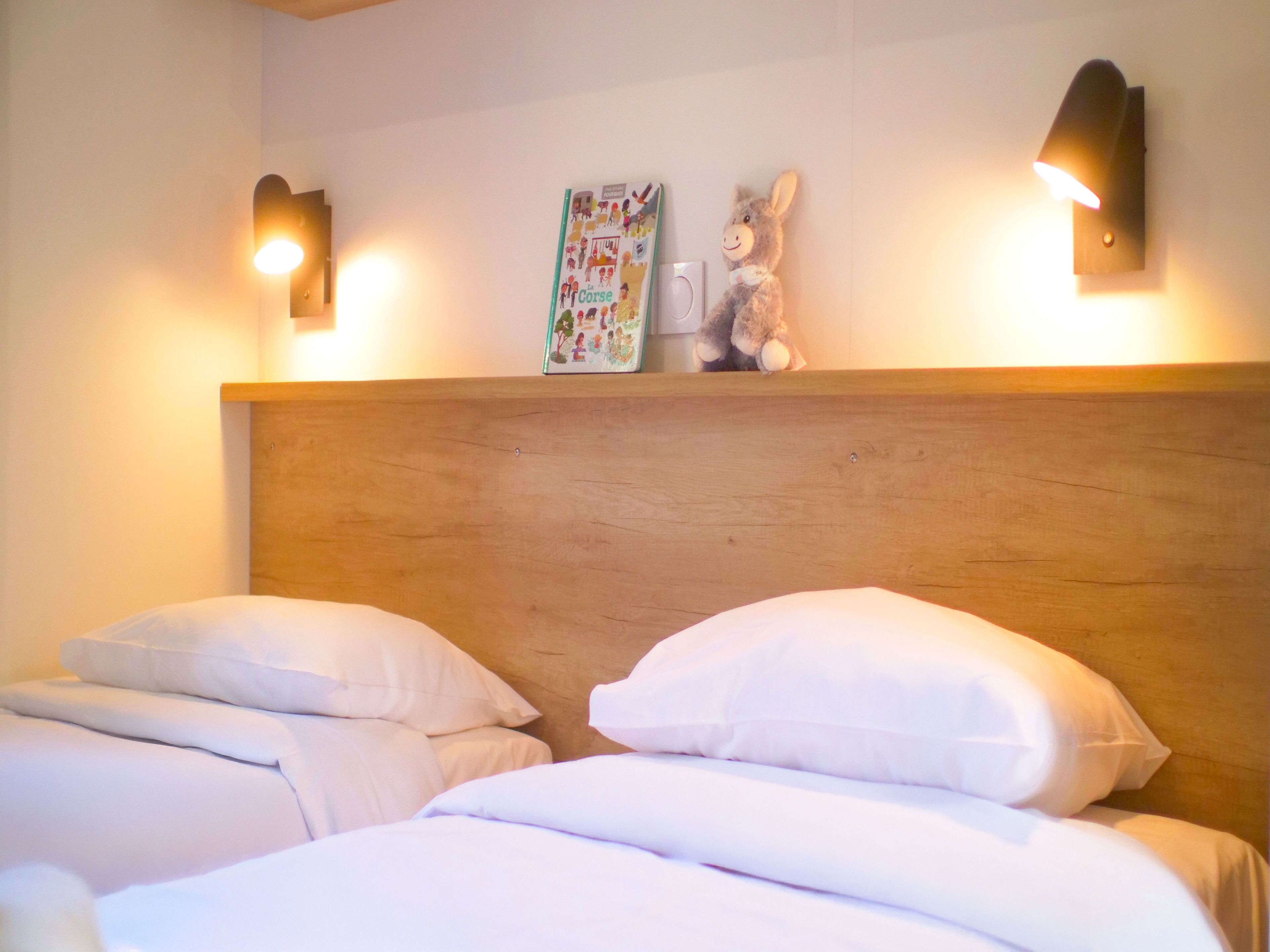 Habitación de hotel con dos camas, cabecero de madera, luces fijadas a la pared y un libro infantil en la pared.