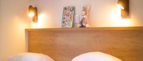 Habitación de hotel con dos camas, cabecero de madera, luces fijadas a la pared y un libro infantil en la pared.