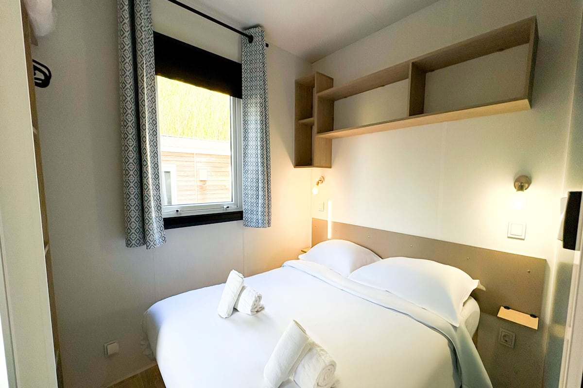 A neatly made bed with white linens and two rolled towels, a window with patterned curtains, and a wall-mounted shelf above the bed.