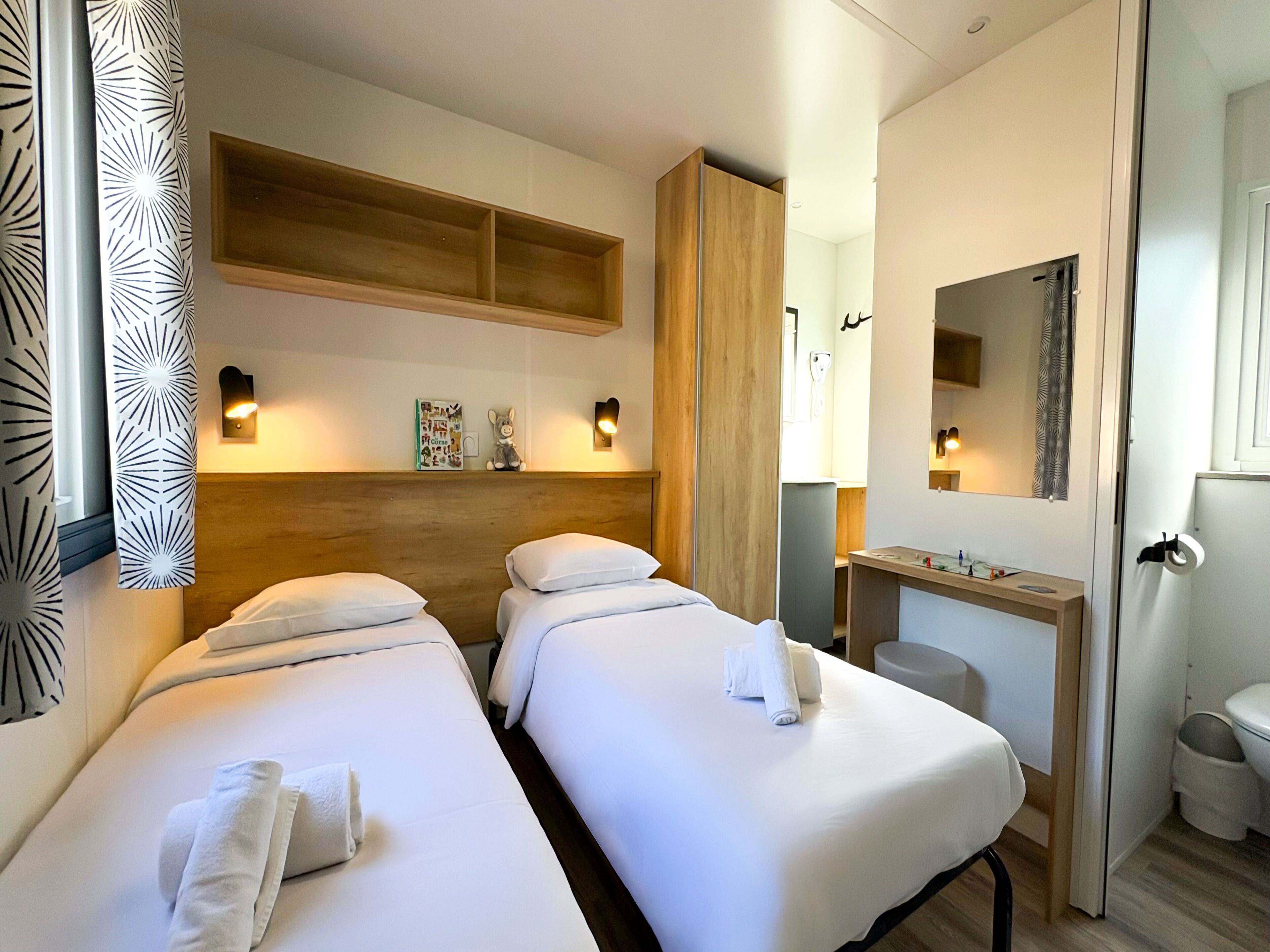 Habitación de hotel con dos camas, un cabecero de madera, un escritorio pequeño con espejo y un baño con inodoro y lavabo.
