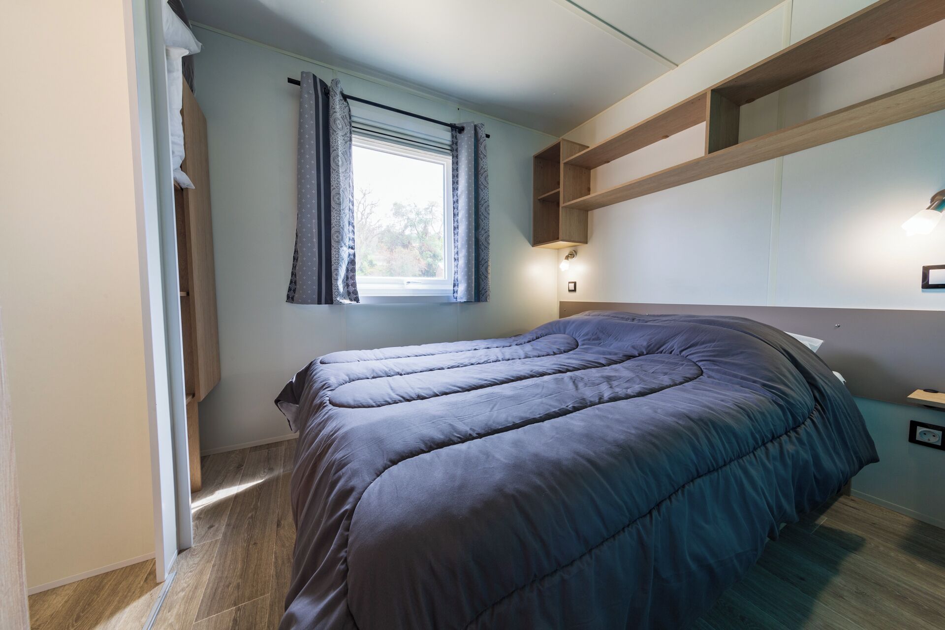 Een slaapkamer met een bed, een raam met gordijnen, een nachtkastje en een wandplank.