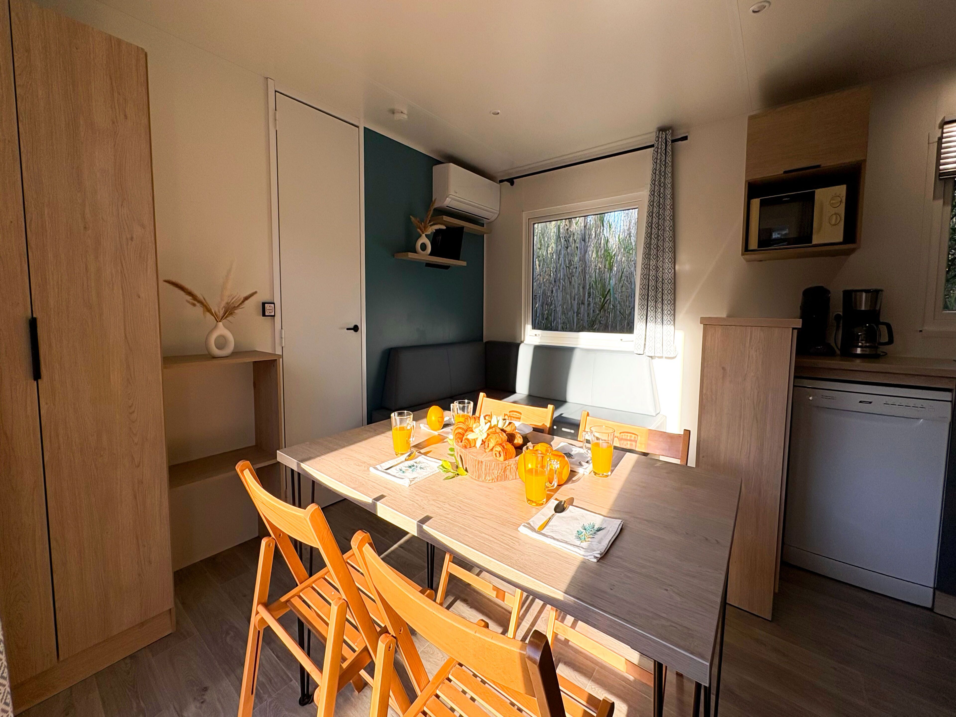 Une cuisine compacte avec un coin repas, des meubles en bois, un four encastré et une fenêtre donnant sur un espace verdoyant.