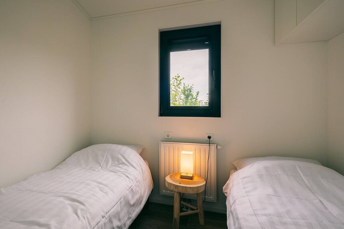 A small, sparsely furnished room with two single beds, a small table, and a window with a view of greenery.