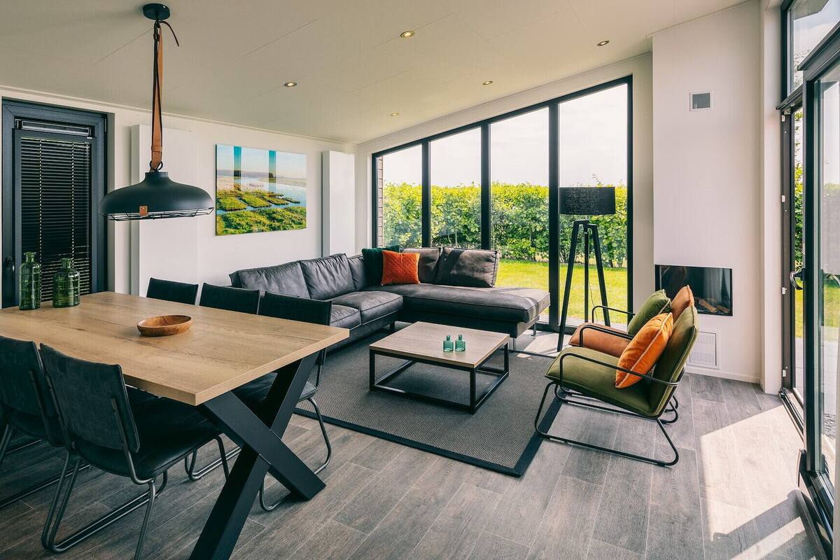 A modern living room with a wooden dining table, black leather sofa, and a large window offering a view of greenery.
