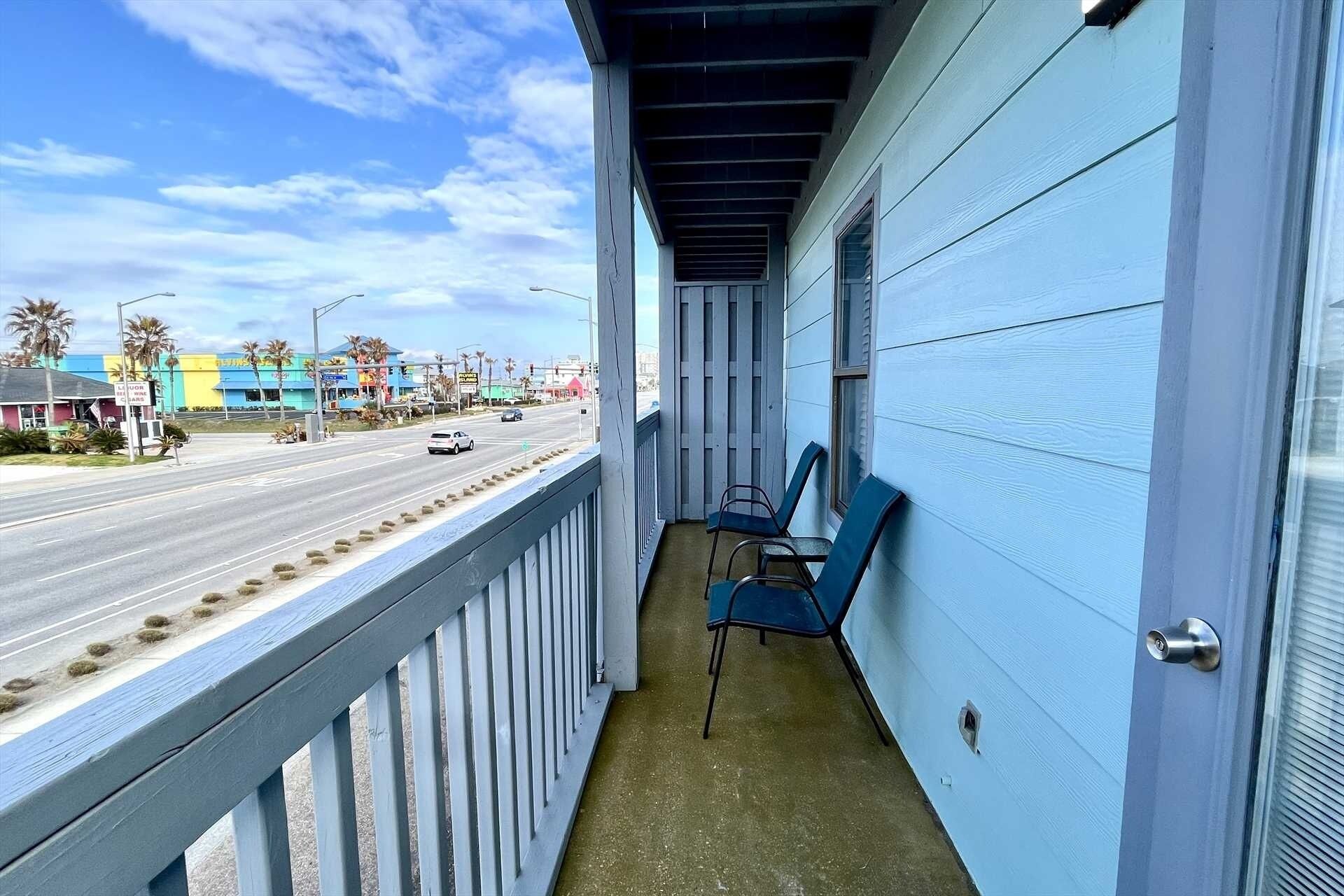 Een balkon met twee stoelen, uitzicht op een straat met palmbomen en gebouwen, en een heldere lucht.