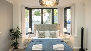 A modern bedroom with a large bed, two bedside tables with lamps, a potted plant, and a view through a large window.