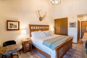 A bedroom with a wooden bed, a wall-mounted deer head, a small table with a lamp, and a chair. - 1BD at Trailhead • Top Floor • Mtn Views (Crested Butte)