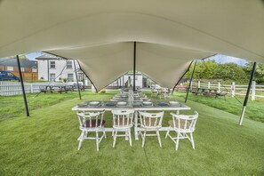 Un espace de restauration en plein air, aménagé sous une tente, avec une longue table et des chaises blanches, entouré d’une clôture en bois peinte en blanc et de verdure.