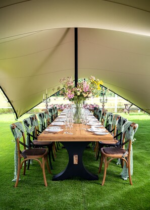 Un área de comedor con carpas, una larga mesa de madera dispuesta para una comida, rodeada de sillas con tela verde.