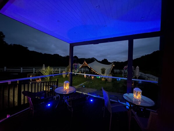 Un patio al aire libre con mesas y sillas, vista de un área decorada al fondo y techo de madera.