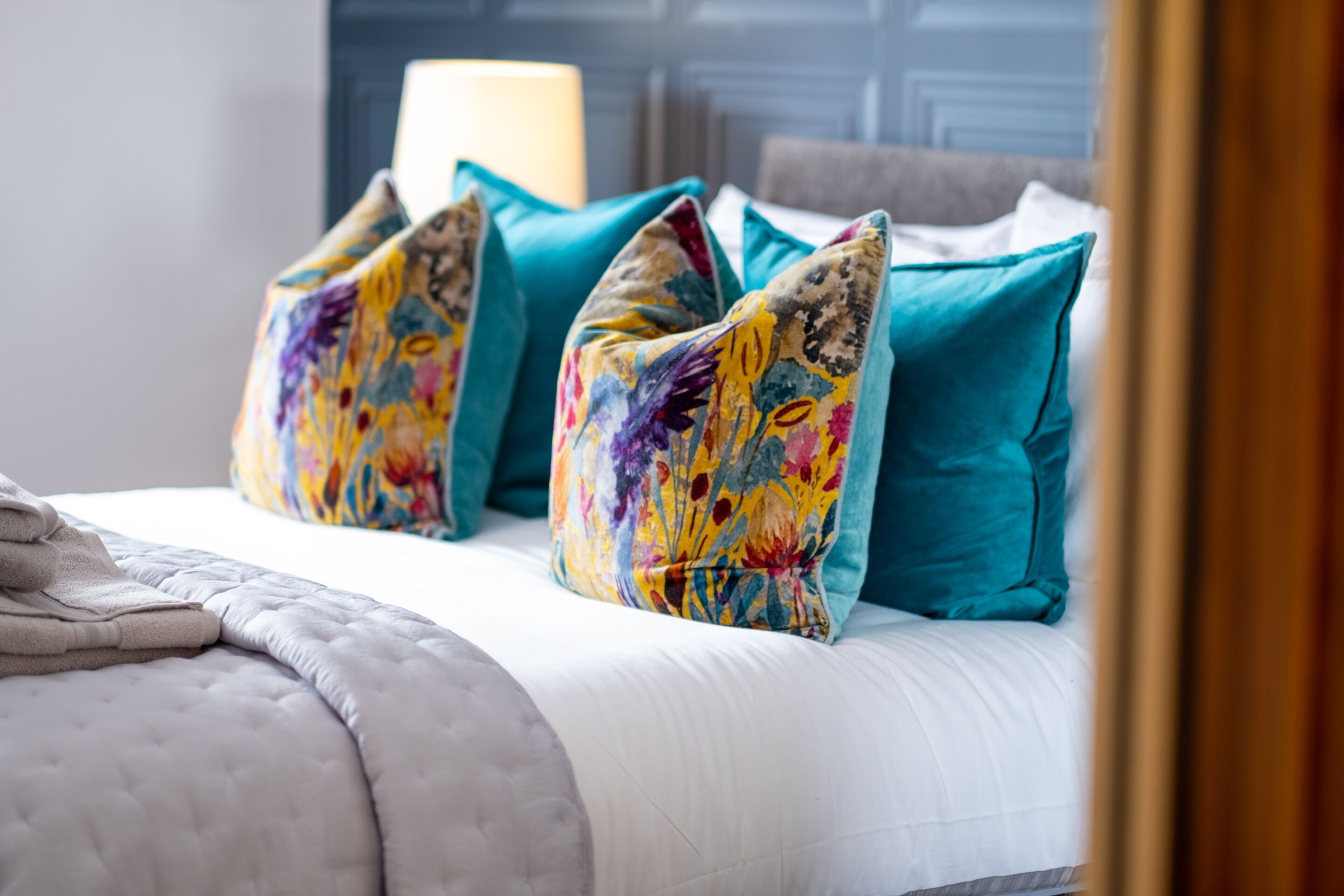 Un lit avec une couette blanche, des oreillers turquoise et un coussin aux motifs floraux colorés.