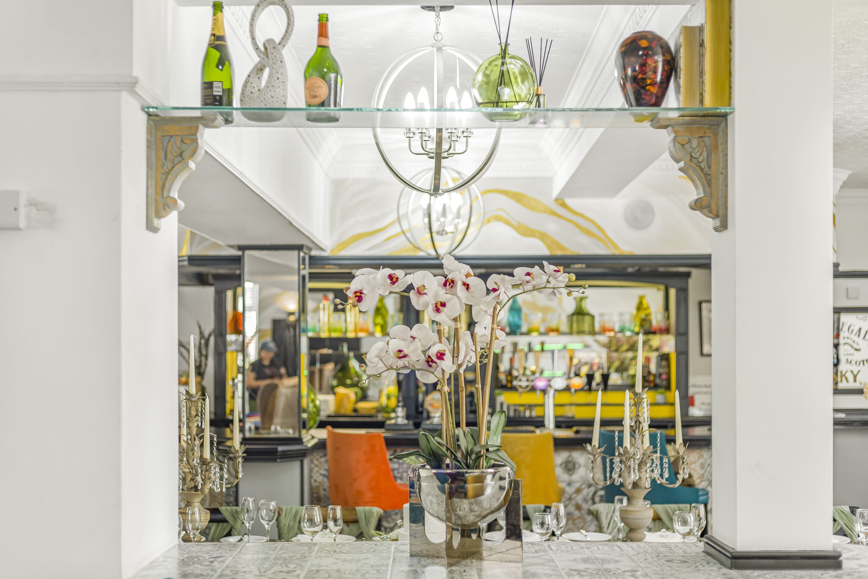 Eine Bar mit einer Glaskonsole, die Flaschen präsentiert, einer Kristallleuchtreiter und einer Topfpflanze mit Orchideen.