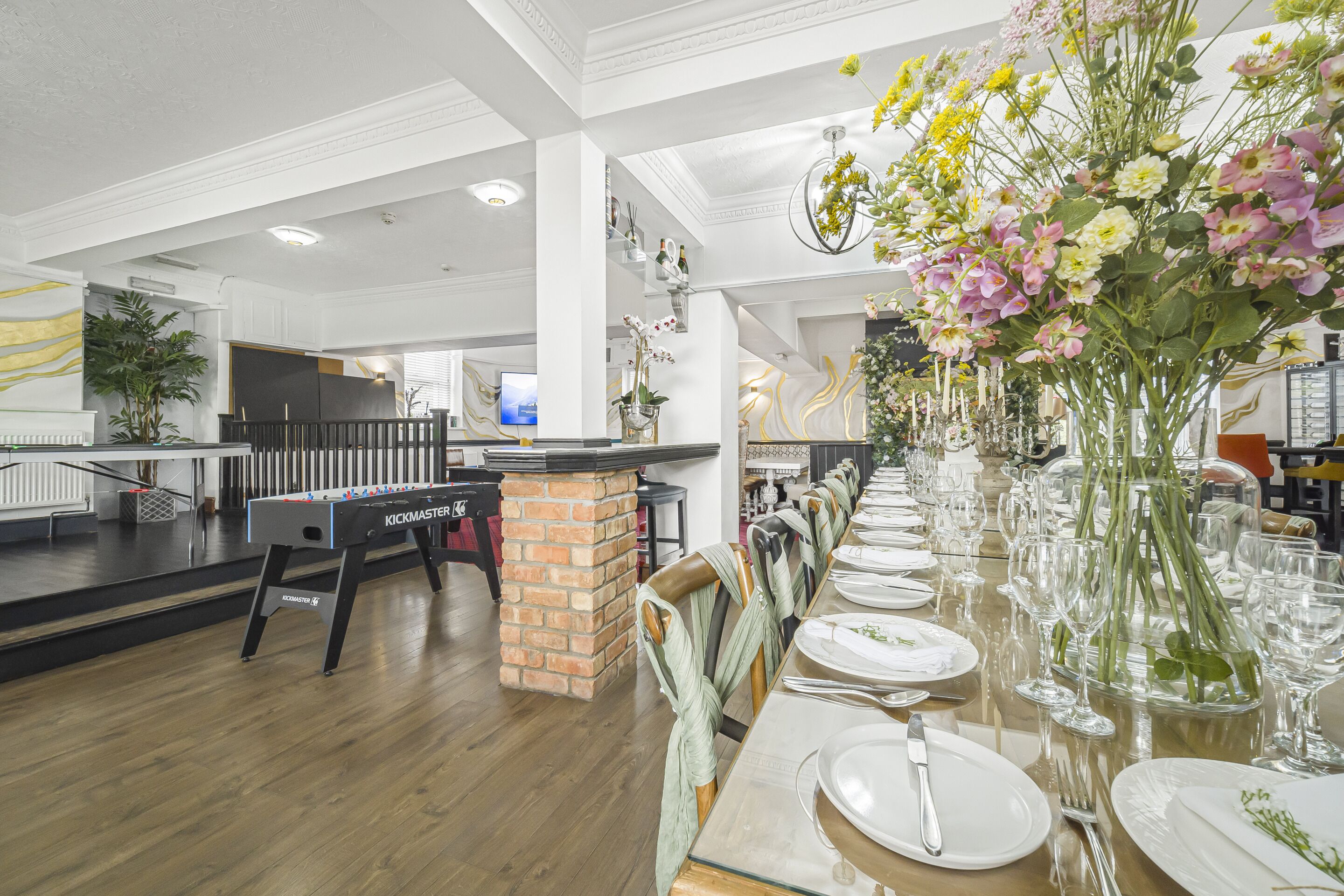 A restaurant with a long dining table set for a meal, a foosball table, and a brick pillar.