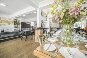Une salle à manger avec une longue table dressée pour un repas, un grand arrangement floral en guise de centre de table, et une table de billard en arrière-plan.