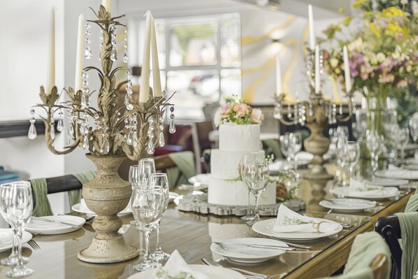 Una larga mesa de comedor dispuesta con copas, platos y una tarta de varios niveles, rodeada de candelabros y arreglos florales.