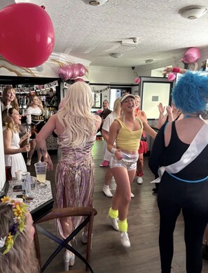 Un grupo de personas bailando en una sala con techo estampado y un área de bar al fondo.