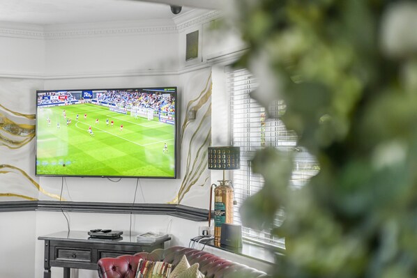 Une pièce comprenant un grand téléviseur à écran plat fixé au mur, sur lequel est diffusé un match de football, un canapé en cuir, une petite table et un élément décoratif mural.
