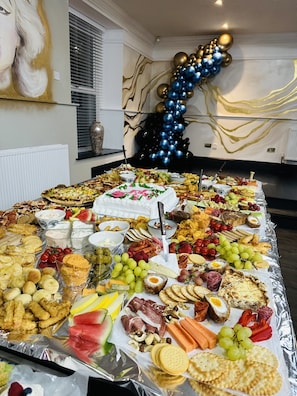 A buffet table with a variety of food items, including fruits, cheeses, and meats, set up in a room with a decorative wall and a window with blinds.