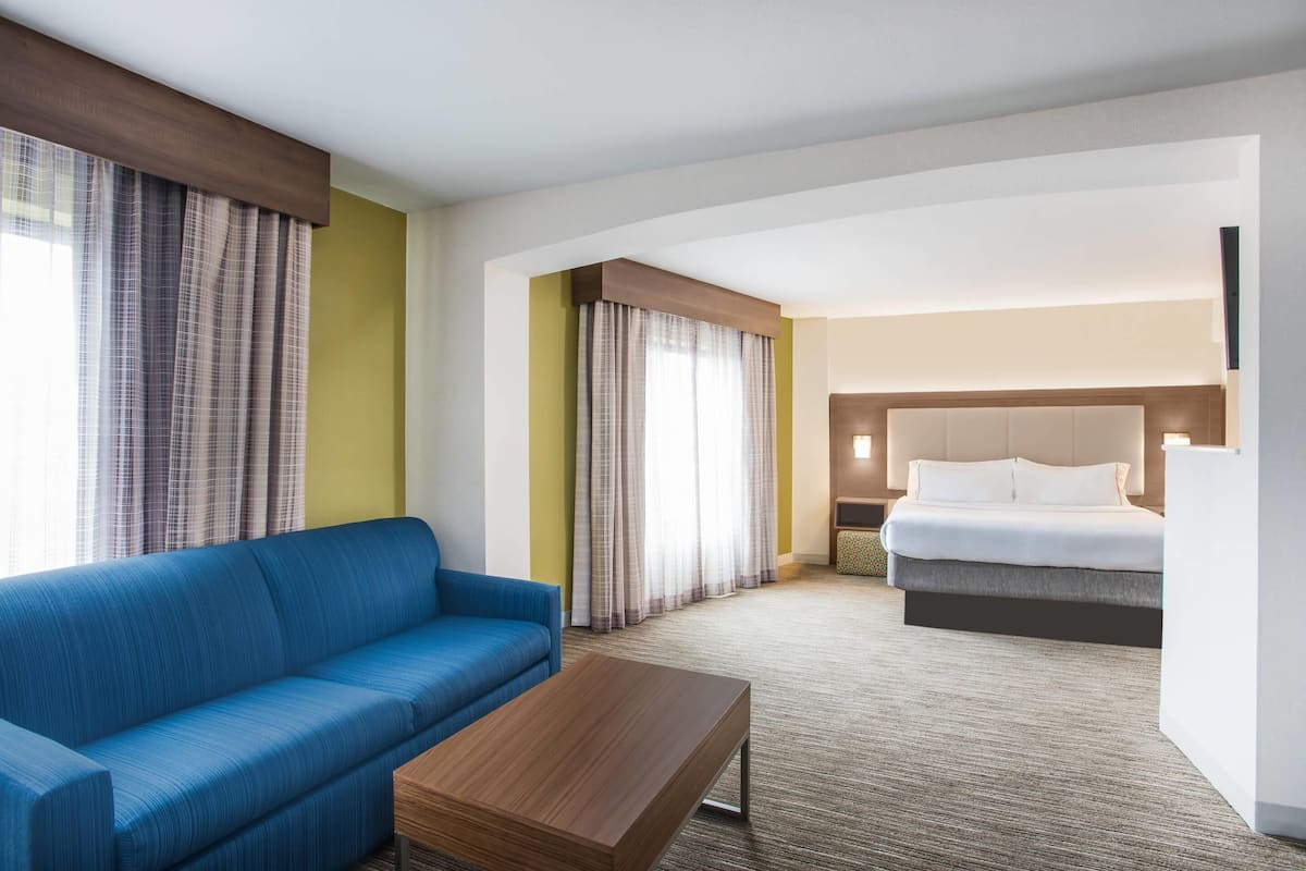 A hotel room with a blue sofa, a wooden coffee table, a bed with a headboard, and curtains on the windows.