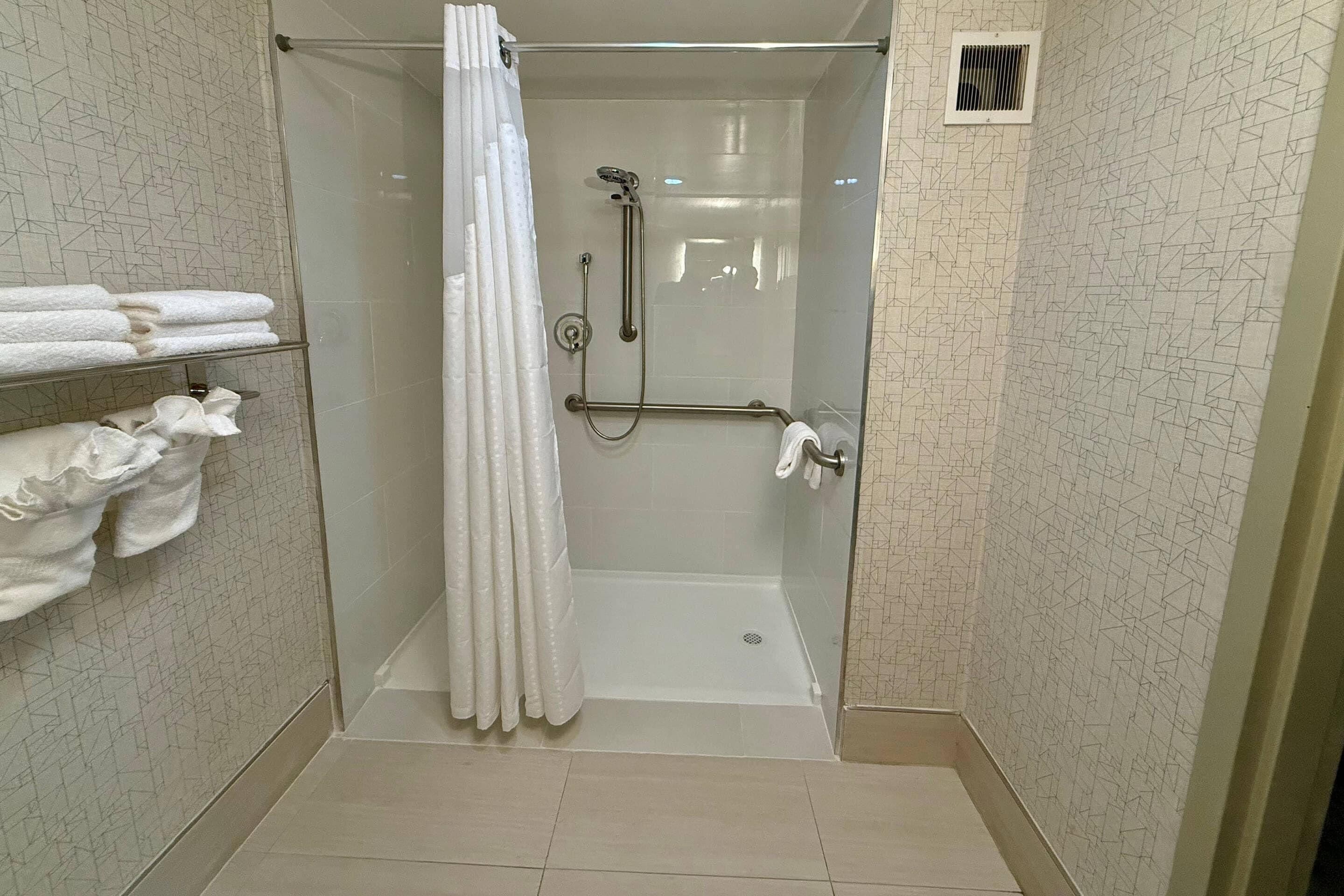 Zona de ducha con mampara de vidrio, cabezal de ducha y ducha de mano, perchero con toallas dobladas y un respiradero en la pared.