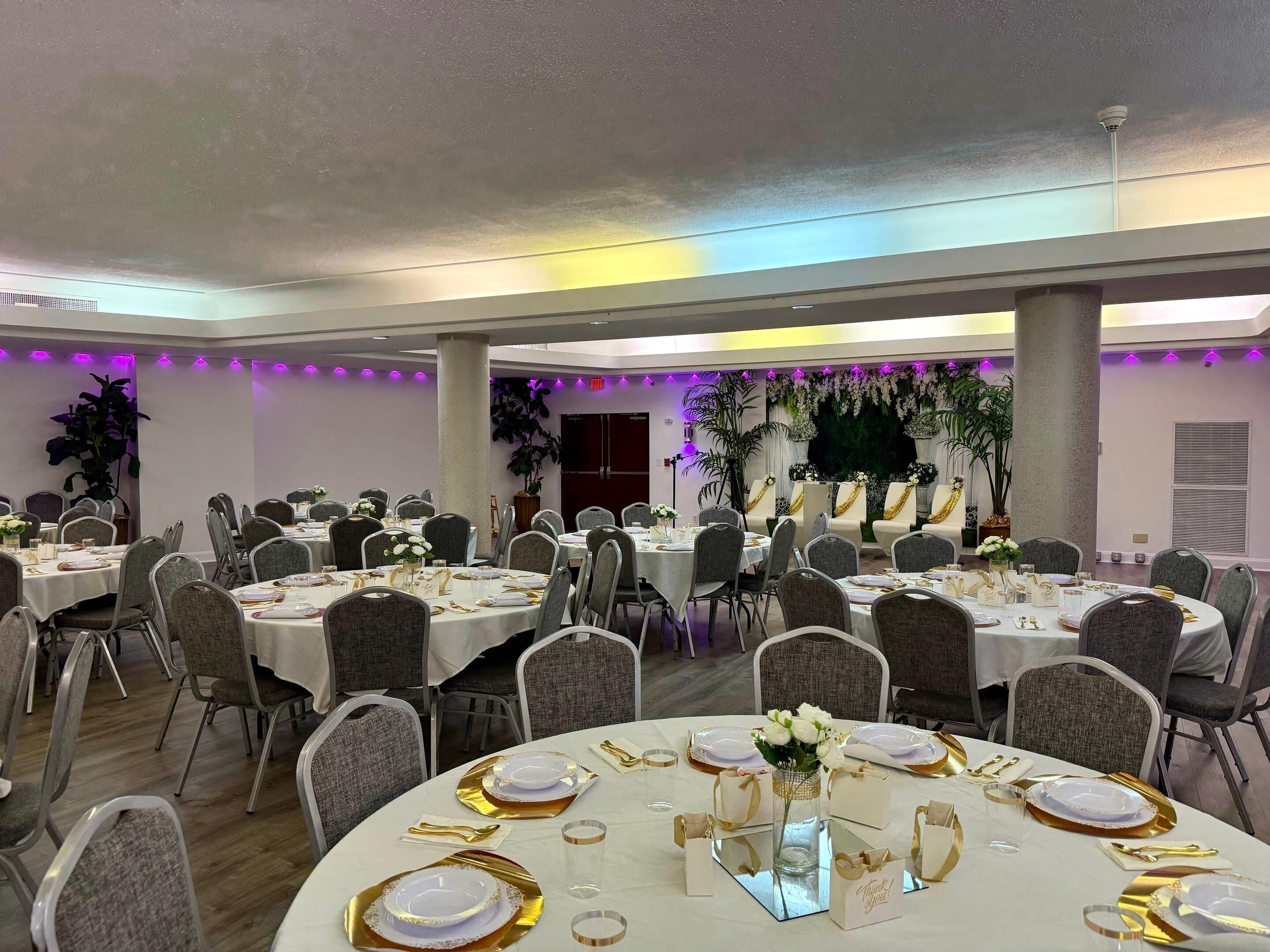 A banquet hall with round tables set for dining, featuring gold and white tableware, and surrounded by grey chairs.