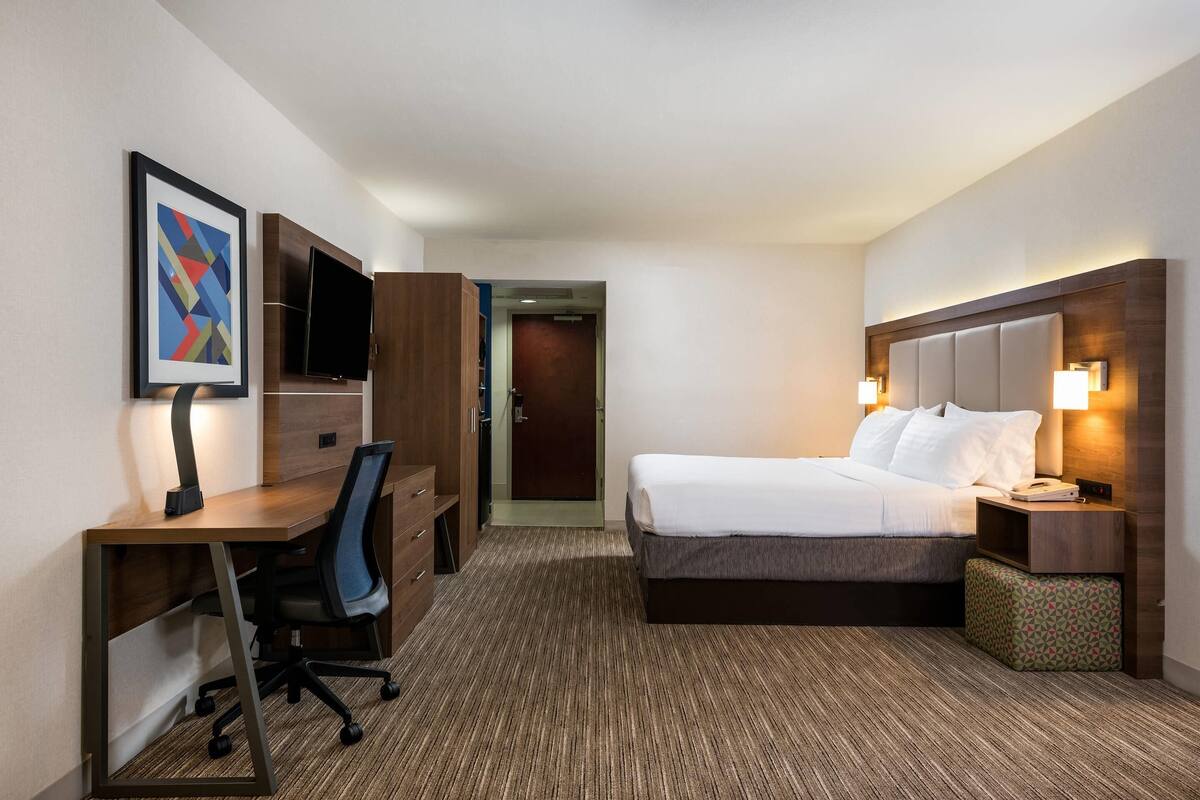 A hotel room with a bed, desk, chair, TV, and a small bedside table with a lamp.