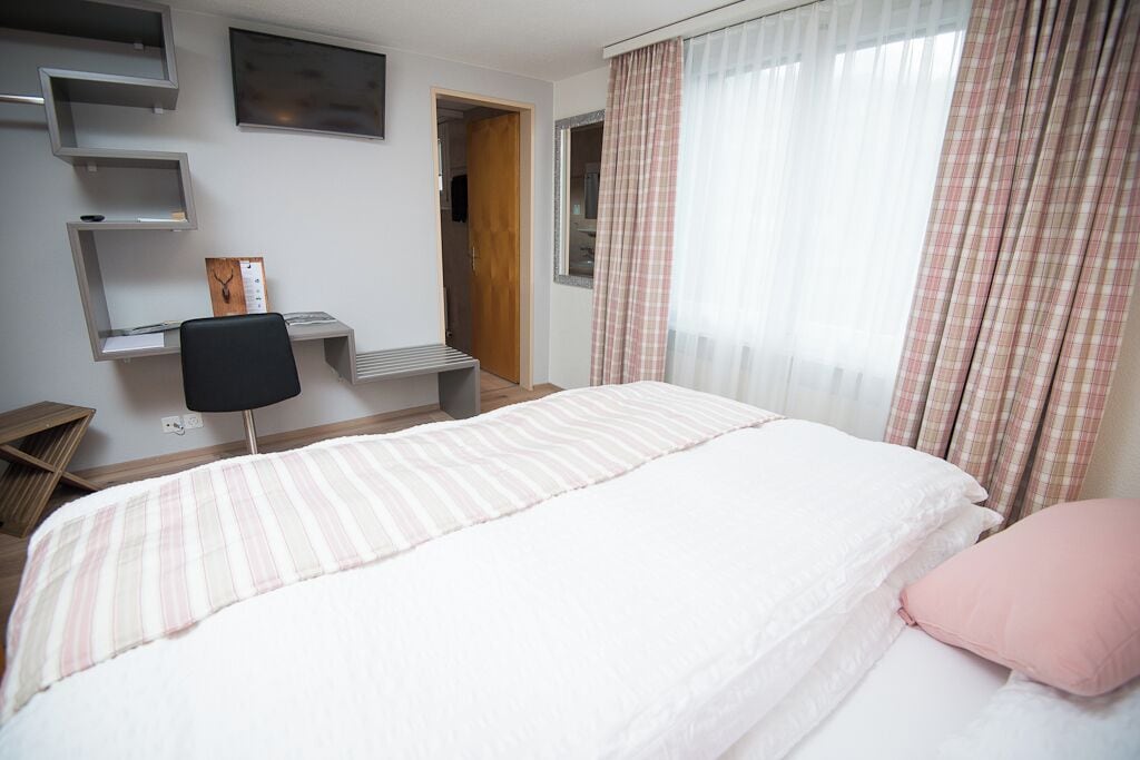 Ein Schlafzimmer mit einem Bett, einem Schreibtisch und einem Stuhl. Ein Fenster mit Vorhängen und eine Tür zu einem anderen Raum.