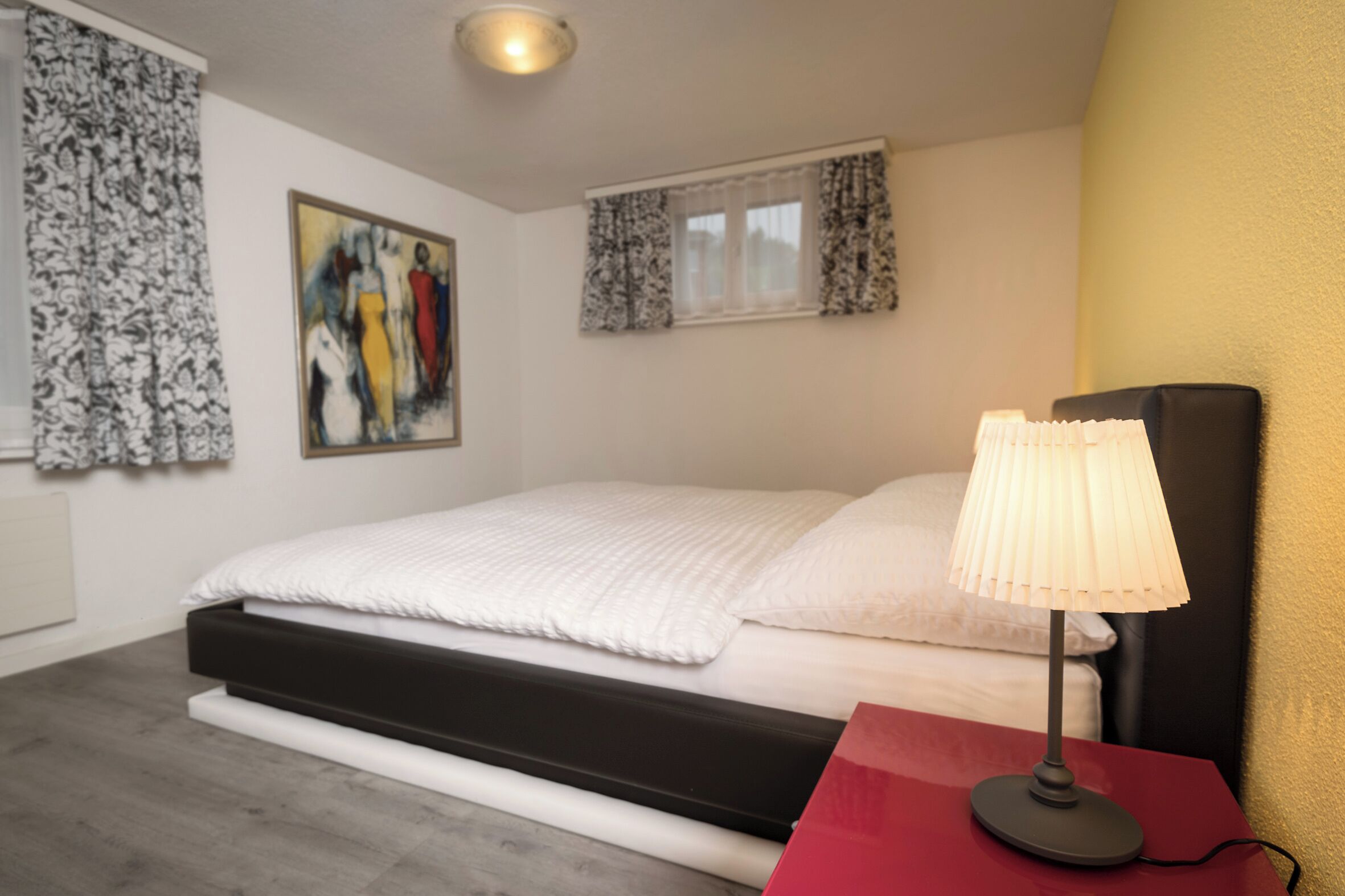 A bedroom with a bed, a red nightstand, a lamp, and a painting on the wall.