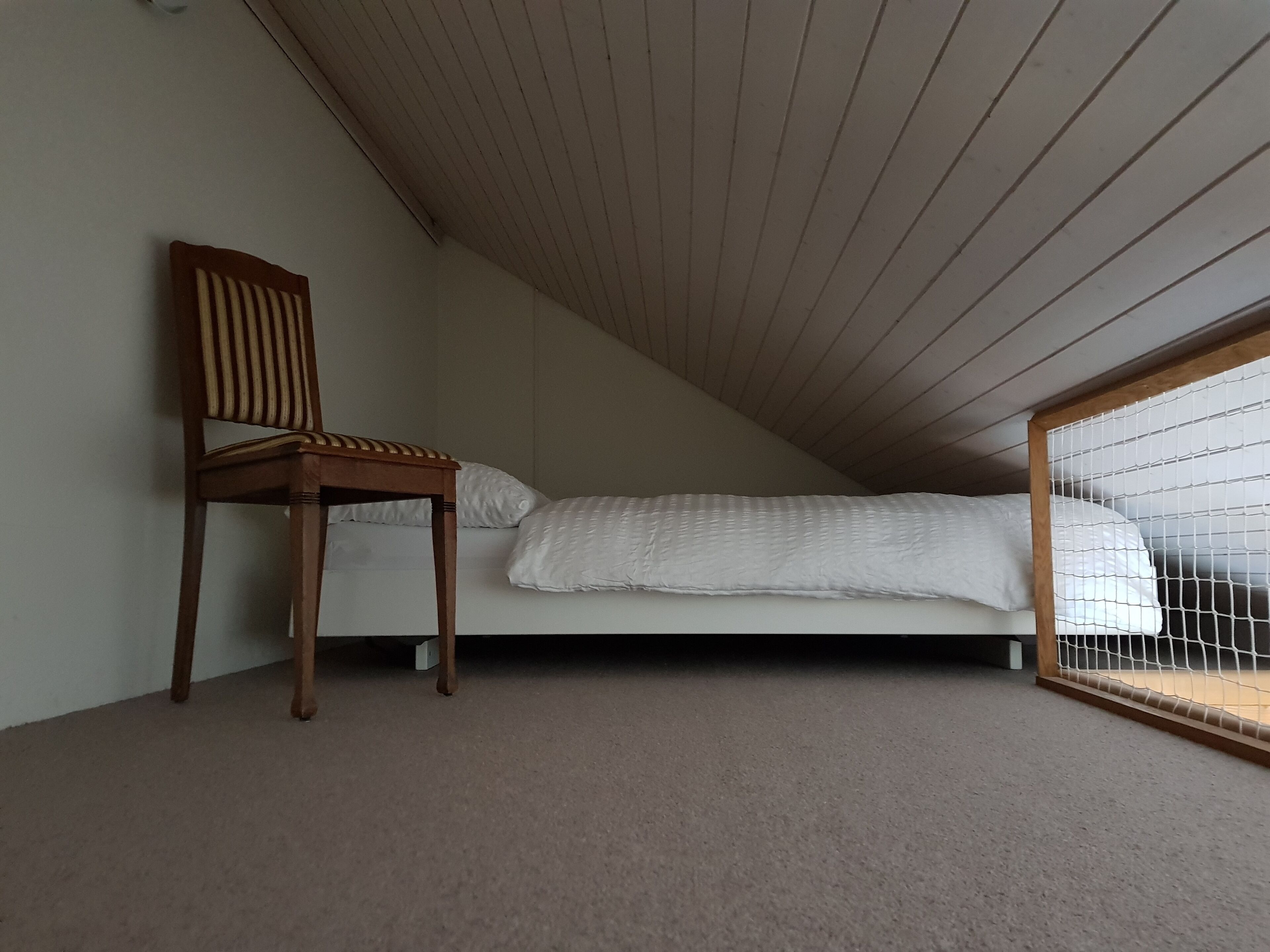 A room with a sloped ceiling, a single bed with white bedding, a wooden chair, and a window with a white blind.