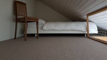 A room with a sloped ceiling, a single bed with white bedding, a wooden chair, and a window with a white blind.