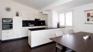 A modern kitchen with white cabinetry, a black countertop, and a dining area with a wooden table. - Panoramarestaurant Hotel Roggerli  (Hergiswil)