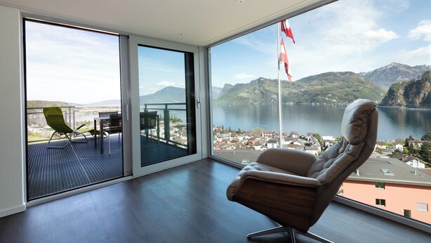 A spacious room with a large sliding glass door leading to a balcony overlooking a lake and mountains. - Panoramarestaurant Hotel Roggerli  (Hergiswil)