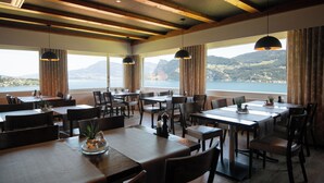 A restaurant with multiple tables and chairs, large windows with a view of mountains and water, and pendant lights. - Panoramarestaurant Hotel Roggerli  (Hergiswil)
