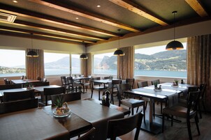 Un restaurante con varias mesas y sillas, amplias ventanas que ofrecen vistas a montañas y agua, y lámparas colgantes.